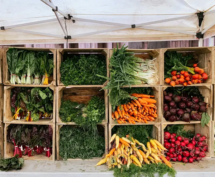 Wooden crates of vegetables stacked under a canopy: greens, carrots, peppers and cabbages.
