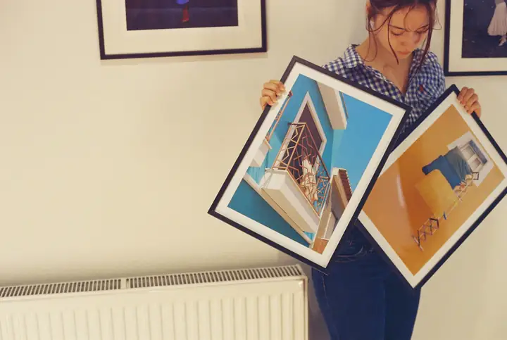 A woman holding up two framed colour prints against a wall hung with more.