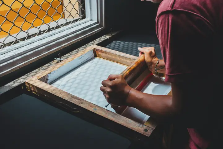 A printer holding a wooden screen frame up to the light of a window.