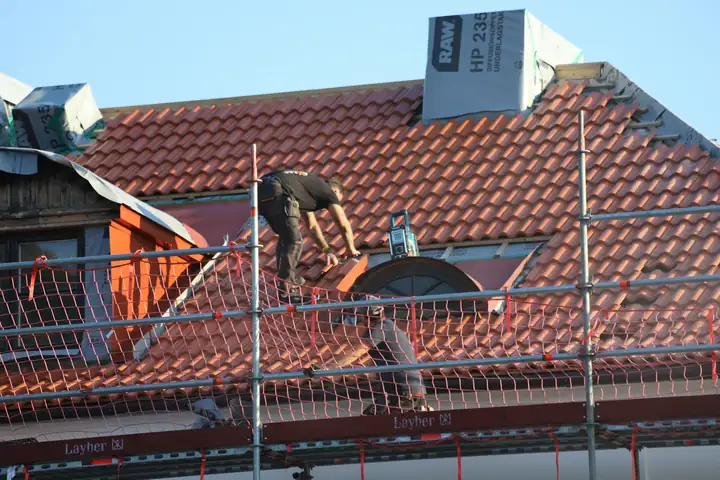 A roofer laying red clay tiles behind scaffolding and orange safety netting.